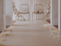 Festlich dekorierter Außenbereich für eine Hochzeit mit rosa Teppich in der Mitte, rechts und links Sitzgruppen aus Rattan, große Blumengestecke in weißen Töpfen am Teppichrand, Sonnenschirme und ein Pavillon am Ende des Teppichs, im Hintergrund das Meer und ein Segelboot.