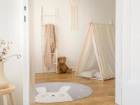 Helles, kinderfreundliches Zimmer mit Holzfußboden, einer dekorativen Holzleiter mit beiger Decke, einem Stoffbären, einem kleinen Kinderzelt mit hellen Kissen, rundem Teppich mit Hasengesicht-Motiv und neutraler Wanddekoration.
