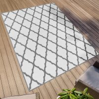 Rechteckiger Outdoor-Teppich mit weißem Hintergrund und grauem, rautenförmigem Gittermuster auf einer Holzterrasse, im Vordergrund ein Teil einer Sitzgruppe und eine Pflanze.