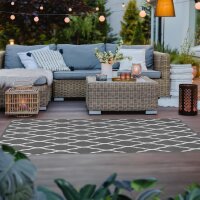 Gemütlich eingerichtete Terrasse mit einem Ecksofa aus Rattan mit hellgrauen Kissen, Couchtisch aus Rattan, dekoriert mit Blumen und Kerzen, stimmungsvolle Beleuchtung durch Lichterkette und Laternen, moderner Teppich mit weißem Rautenmuster auf dunklem Boden, umgeben von Pflanzen.