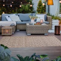 Gemütliche Outdoor-Sitzecke mit einem Ecksofa aus Rattan und hellgrauen Kissen, dekoriert mit weißen und gemusterten Kissen sowie einem Fell. Vor dem Sofa steht ein Rattan-Tisch, darauf ein Tablett mit Kerzen und eine Vase mit weißen Blumen. Die Szene ist stimmungsvoll beleuchtet mit Lichterketten, einer Stehlampe und Kerzen in Laternen. Im Vordergrund liegt ein Teppich mit Zickzack-Muster, im Hintergrund sind Pflanzen und Garten sichtbar.