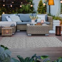 Gemütlich eingerichtete Terrasse mit einem grauen Rattansofa, hellen Kissen und Decken, flachem Rattantisch mit Blumen und Kerzen sowie dekorativer Hängelampe und Lichterkette im Hintergrund, Teppich mit hellem Zickzackmuster und viel Grünpflanzen im Vordergrund.