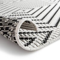 Nahaufnahme eines teilweise aufgerollten Teppichs mit schwarz-weißem geometrischem Muster auf weißem Hintergrund. Die Struktur des gewebten Materials ist deutlich sichtbar.