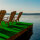 Zwei hölzerne Liegestühle auf einem Schiffdeck mit grünem Bodenbelag, dazwischen ein kleiner Tisch mit drei bunten Cocktails, im Hintergrund Sonnenuntergang über dem Meer und bewölkter Himmel.