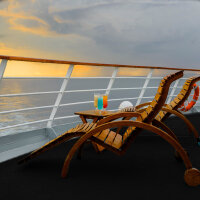 Zwei leere Holzliegestühle stehen auf dem Deck eines Kreuzfahrtschiffs, dazwischen ein kleiner Holztisch mit drei bunten Cocktails, im Hintergrund ist das Meer und ein Sonnenuntergang mit bewölktem Himmel zu sehen.