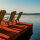 Zwei leere Holzliegestühle stehen auf dem Deck eines Schiffs, daneben ein kleiner Tisch mit drei bunten Cocktails. Im Hintergrund ist das Meer zu sehen, am Horizont geht die Sonne unter und es sind Wolken am Himmel.