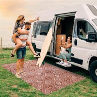 Drei Erwachsene genießen gemeinsam die Zeit vor einem Campervan: Ein Mann trägt eine Frau huckepack, beide lachen. Neben ihnen lehnt ein Surfbrett an dem Fahrzeug. Eine weitere Frau sitzt auf der offenen Türschwelle des Campers, hält ein Handy und ein Getränk und hat eine Katze auf dem Schoß. Unter ihnen liegt ein gemusterter Teppich auf dem Gras, im Hintergrund ist das Meer zu sehen. Das Bild vermittelt eine fröhliche, entspannte Campingatmosphäre bei Sonnenuntergang.