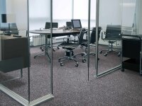 Modernes, helles Besprechungszimmer mit Glastrennwänden, rundem Tisch mit vier schwarzen Bürostühlen auf Rollen, Laptop, Unterlagen und Stiften auf dem Tisch, Teppichboden und natürlichem Tageslicht durch große Fenster.