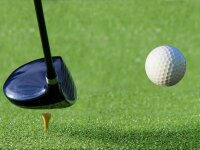 Nahaufnahme eines weißen Golfballs und eines silbernen Putters auf grünem Kunstrasen vor einer grauen Steinwand.