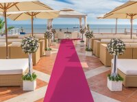 Festlich dekorierter Außenbereich für eine Hochzeit mit rosa Teppich in der Mitte, rechts und links Sitzgruppen aus Rattan, große Blumengestecke in weißen Töpfen am Teppichrand, Sonnenschirme und ein Pavillon am Ende des Teppichs, im Hintergrund das Meer und ein Segelboot.
